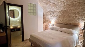 A bedroom with a stone ceiling, a bed with white bedding, a bedside table, and a small bathroom with a mirror and sink. - Santo Stefano Home & Breakfast (Polignano a Mare)