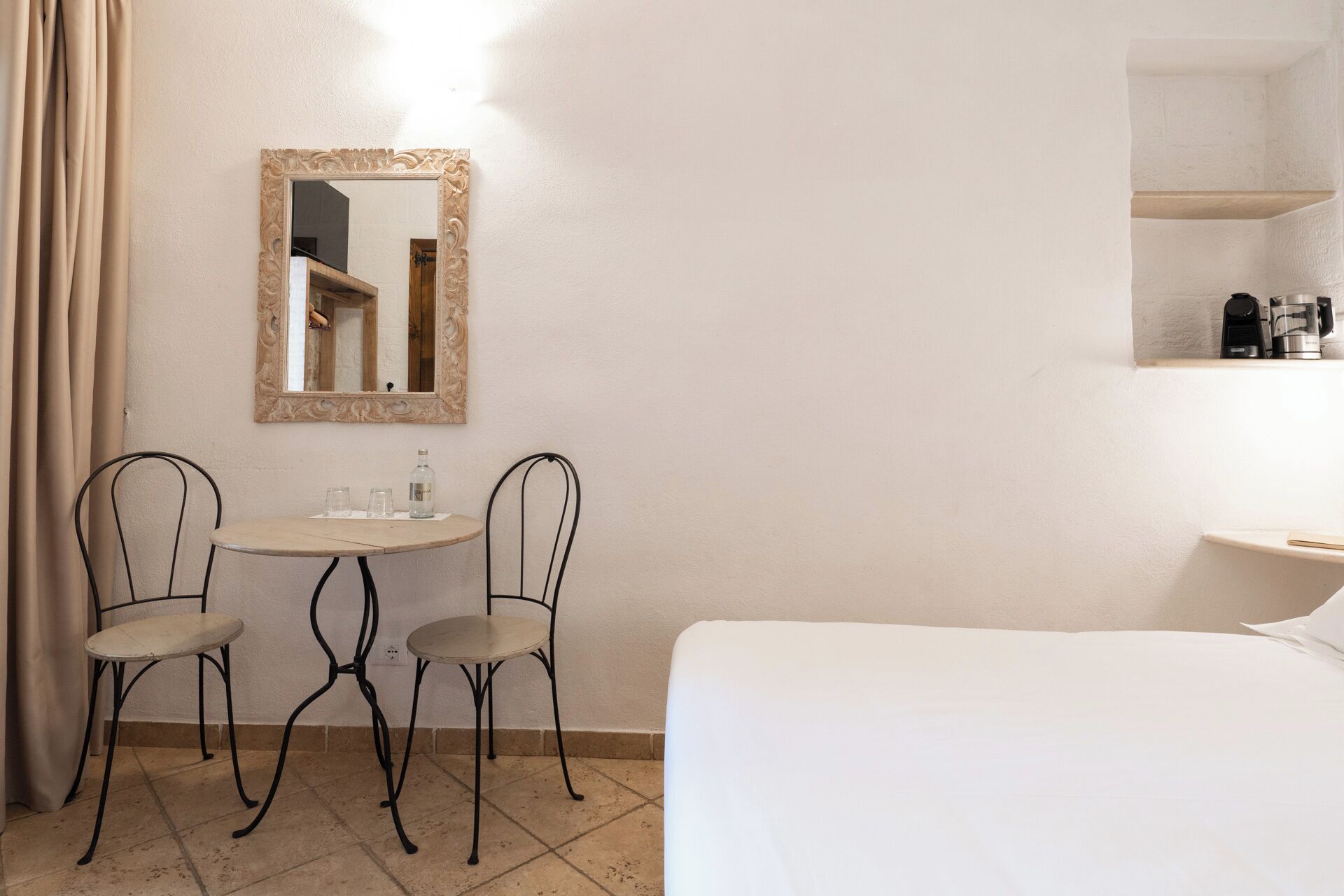 Ein Hotelzimmer mit einem Bett, zwei Stühlen, einem kleinen runden Tisch mit einer Flasche und Gläsern, einem Spiegel und einem Regal mit einer Kaffeemaschine.