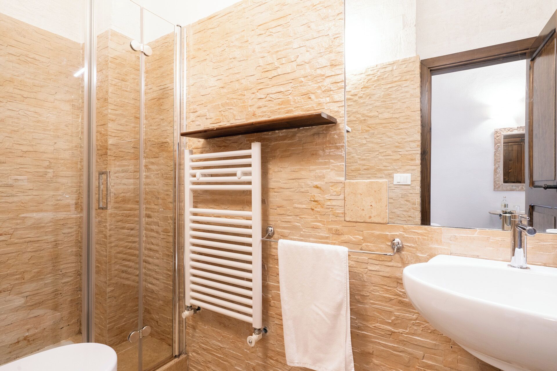 A modern bathroom with a glass shower enclosure, a white towel, a white sink, and a wooden shelf.