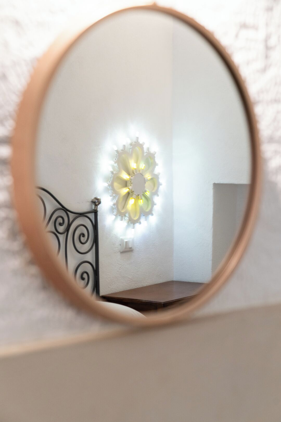 A mirror reflecting a room with a wall-mounted light fixture and a dark-colored bed frame.