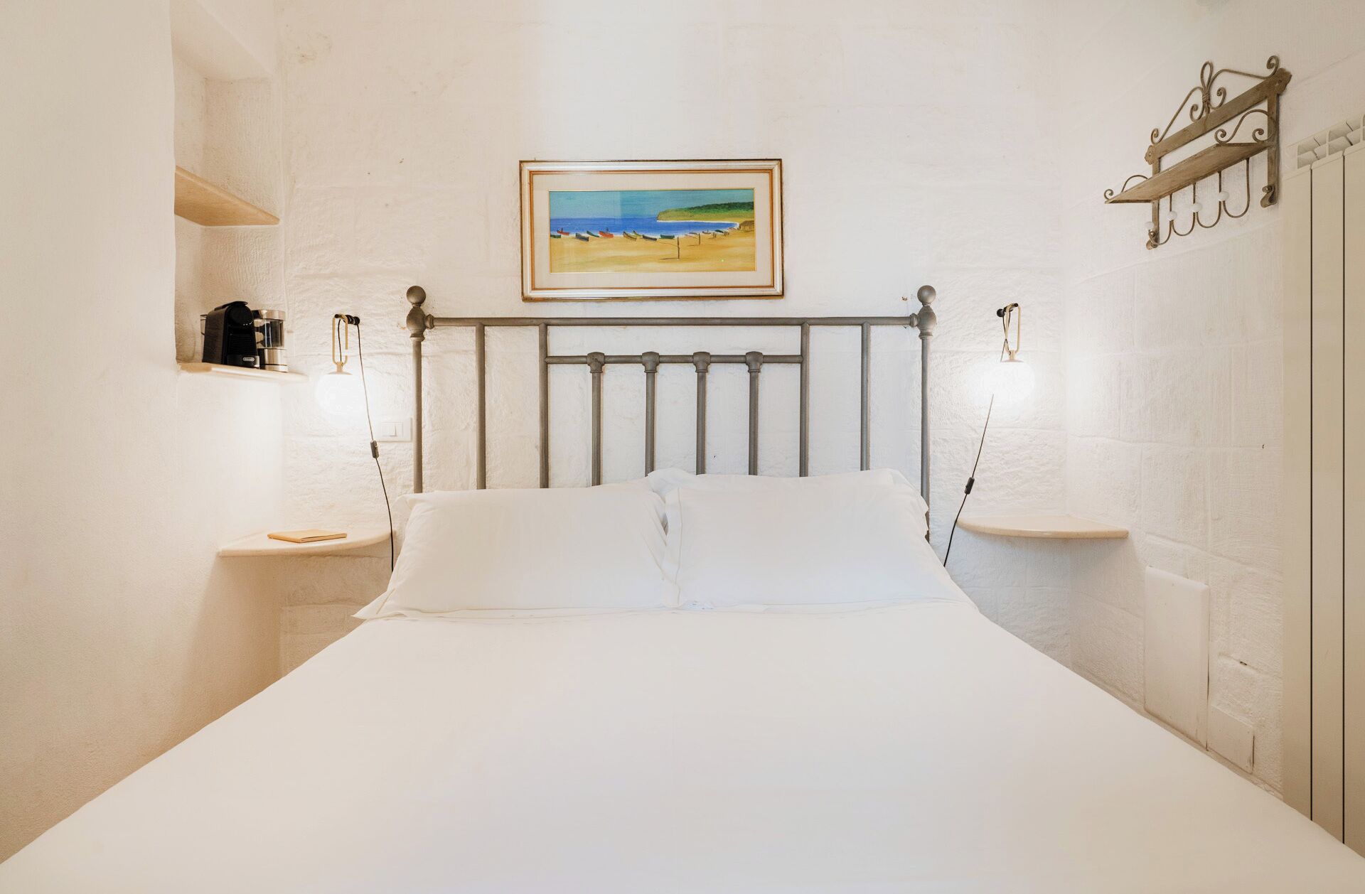 Een slaapkamer met een bed, nachtkastje, een plankje en een foto in een lijst aan de muur, plus een plank met een koffiezetapparaat.