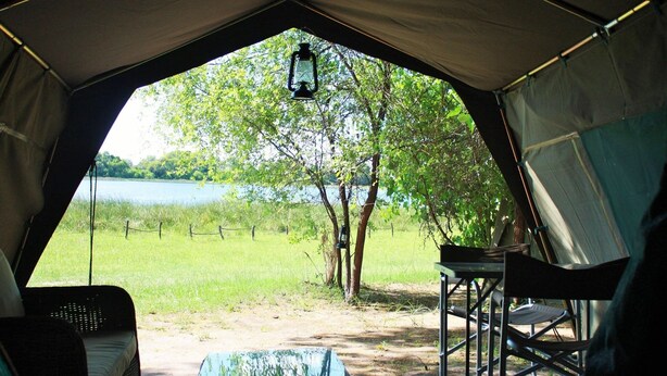 A tent with a view of a lake and trees, a table, and chairs inside. - Hippo Island Okavango Delta (Maun)