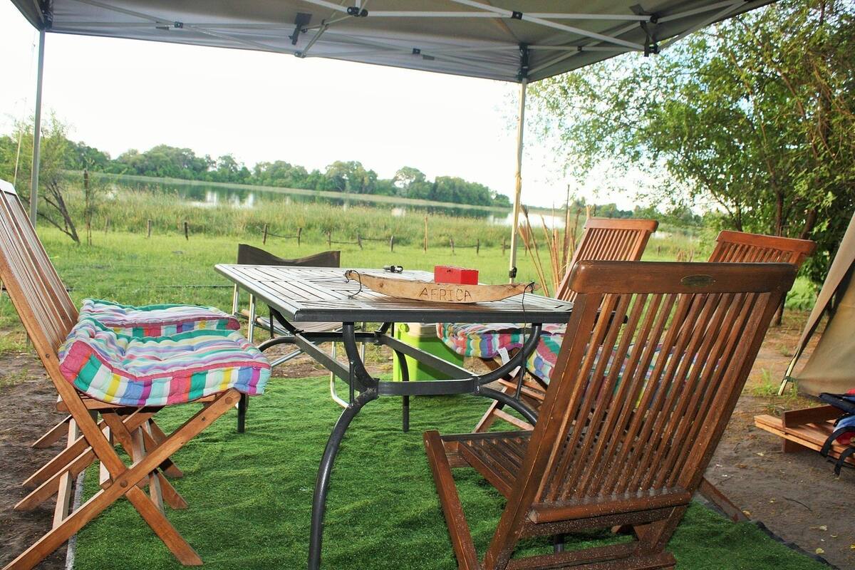 Un área de comedor al aire libre con una mesa, sillas y una carpa, con vista a un lago y vegetación.
