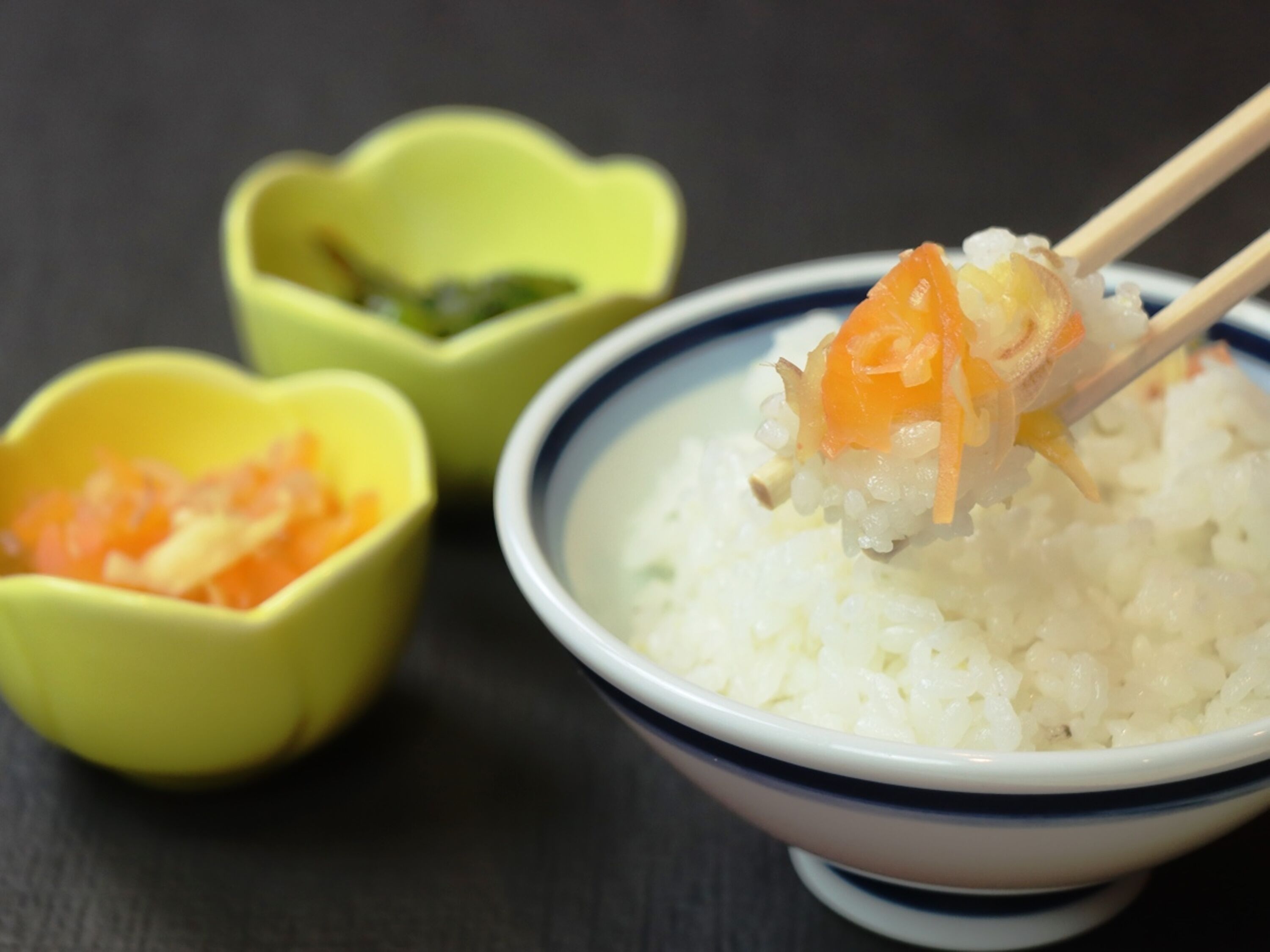 Un tazón de arroz blanco con un trozo de pastel de pescado naranja encima, sostenido por palillos. Hay dos tazones pequeños amarillos con interior verde, uno de los cuales está abierto y contiene una sustancia verde.