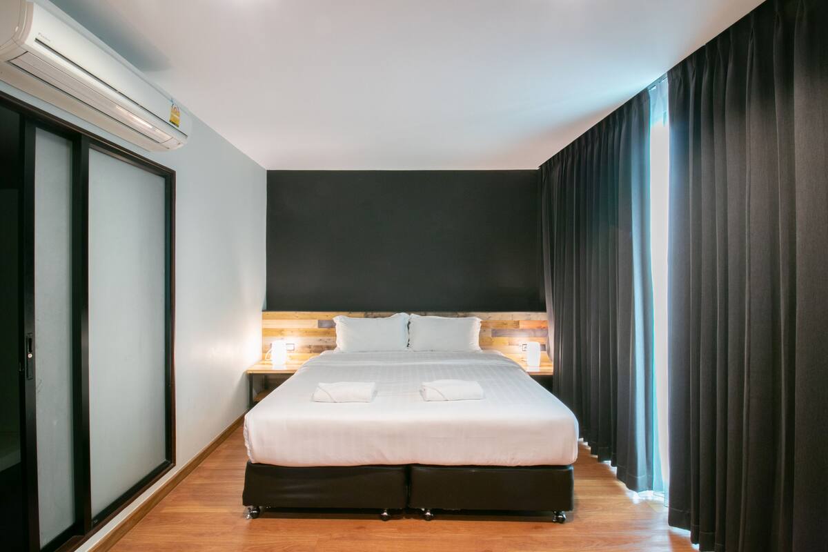 Une chambre d’hôtel avec un grand lit, une tête de lit en bois, une table de chevet et une fenêtre avec des rideaux.