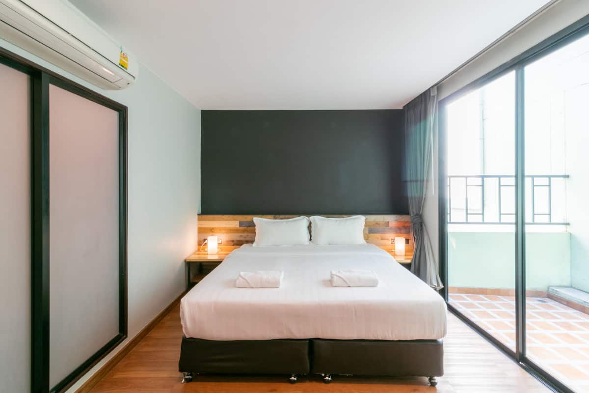 Une chambre d’hôtel avec un grand lit, une tête de lit en bois, une table de chevet avec une lampe et une vue sur le balcon extérieur.