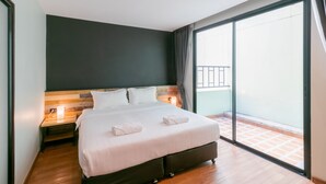 Une chambre d’hôtel avec un grand lit, une table de chevet avec une lampe, une porte-fenêtre coulissante donnant sur un balcon et un mur de couleur sombre.