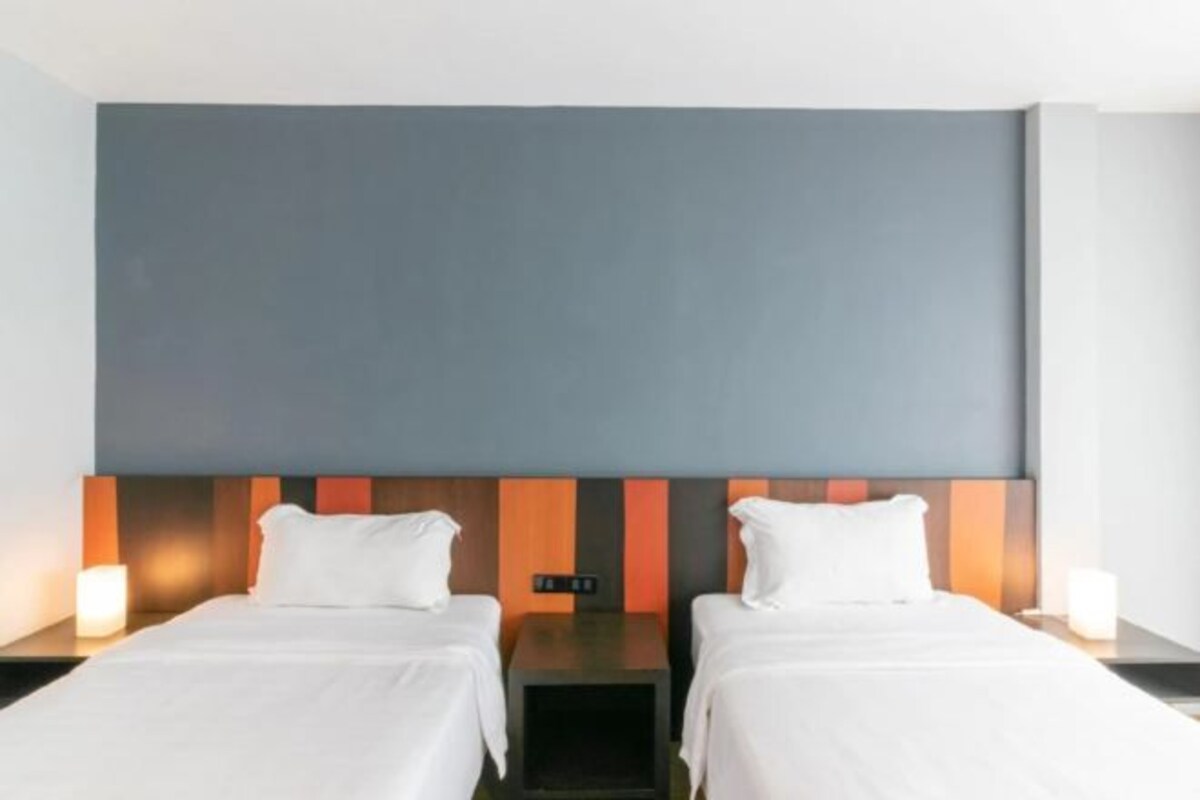 A hotel room with two single beds, a nightstand with a lamp, and a modern headboard with a color-blocked design.