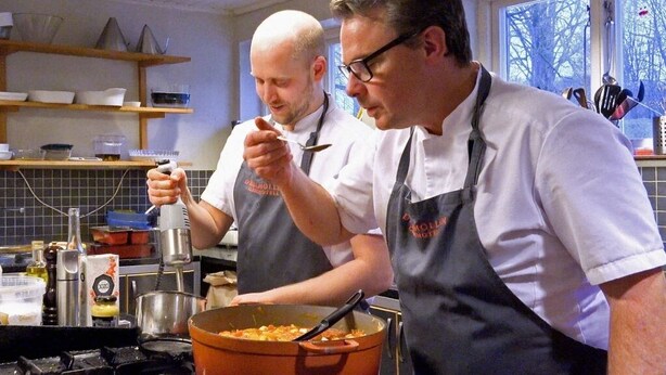 Dos cocineros en una cocina con una olla grande en la estufa, una ventana con vistas a árboles y estantes con utensilios de cocina.