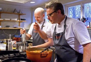 Dos cocineros en una cocina con una olla grande en la estufa, una ventana con vistas a árboles y estantes con utensilios de cocina.
