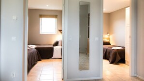 Una habitación con dos camas, una ventana con persianas, un espejo y una puerta que da a otra habitación.