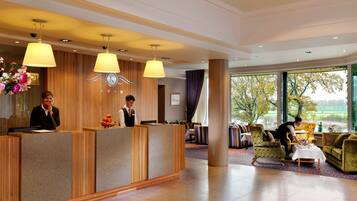 Et hotellets receptionområde med en trædisk, to receptionister, et siddeområde med farverige stole og et stort vindue med udsigt til træer og et mark.