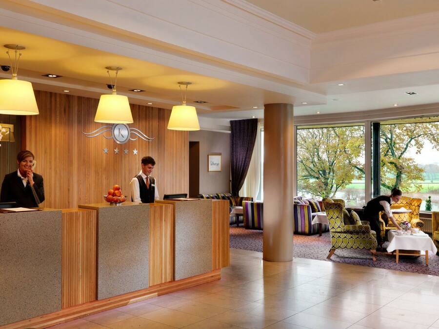 Et hotellets receptionområde med en trædisk, to receptionister, et siddeområde med farverige stole og et stort vindue med udsigt til træer og et mark.