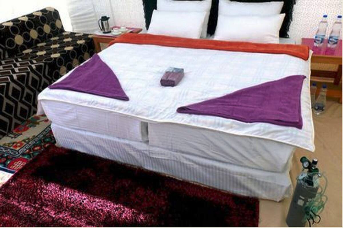 A neatly made bed with a white mattress, a purple blanket, and a red blanket. There is a pillow on the bed, a small table with a kettle, and a water bottle on the floor.