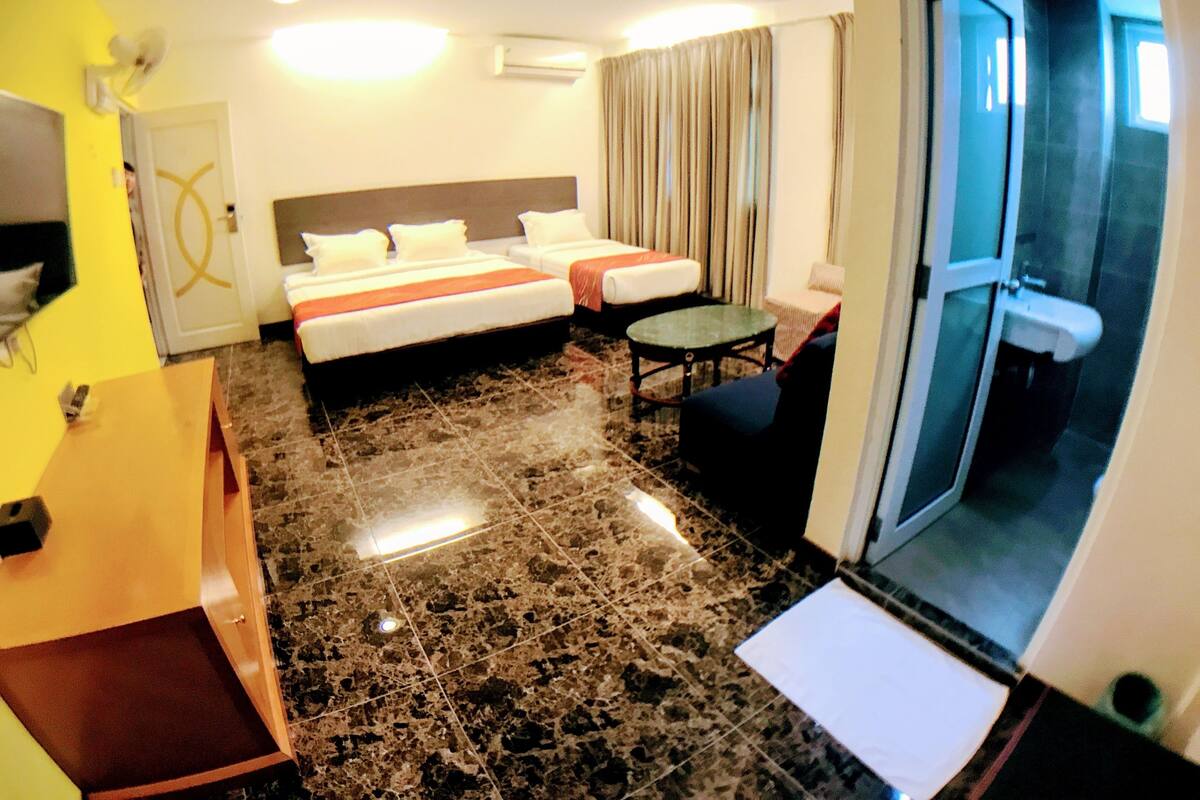 A hotel room with a bed, a desk, a chair, a small table, and a bathroom visible through a glass door.