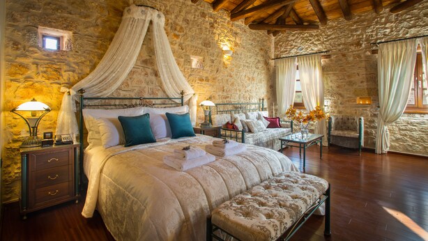 A bedroom with a stone wall, wooden ceiling beams, a four-poster bed, bedside tables with lamps, and a bench. - Mystras Grand Palace Resort & Spa (Sparta)
