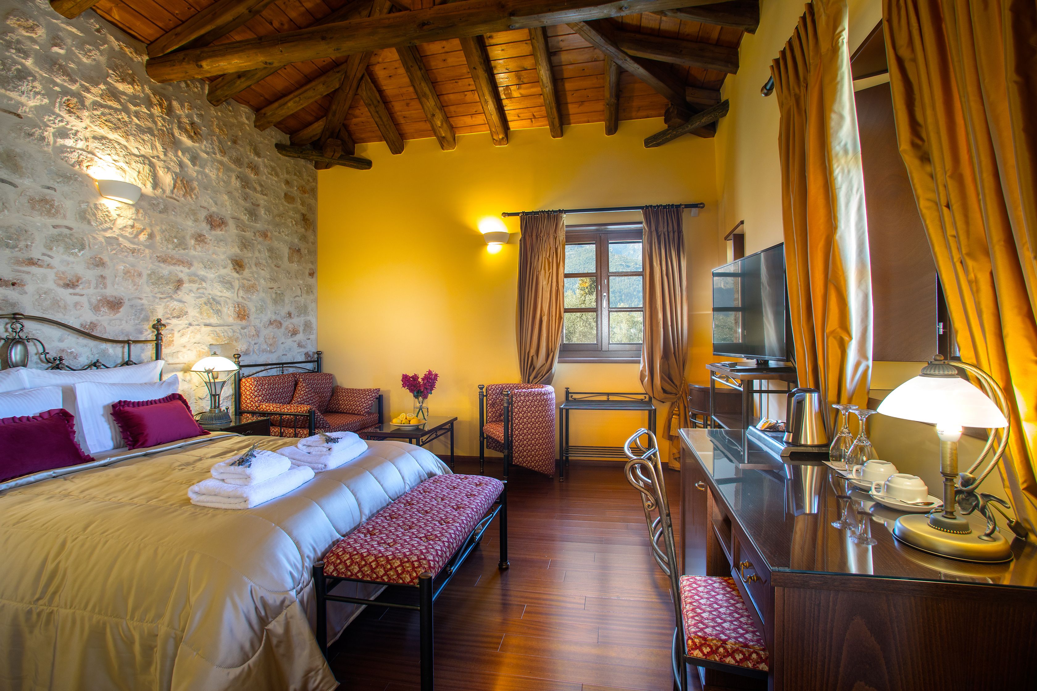 Une chambre d’hôtel avec un mur en pierre, un plafond en bois, un lit avec des oreillers, un banc, un bureau avec une lampe, une chaise, une télévision et une fenêtre avec des rideaux.