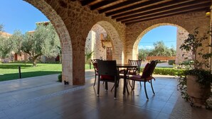 A covered patio with stone arches, a table with chairs, and a view of a courtyard with trees and buildings. - Mystras Grand Palace Resort & Spa (Sparta)