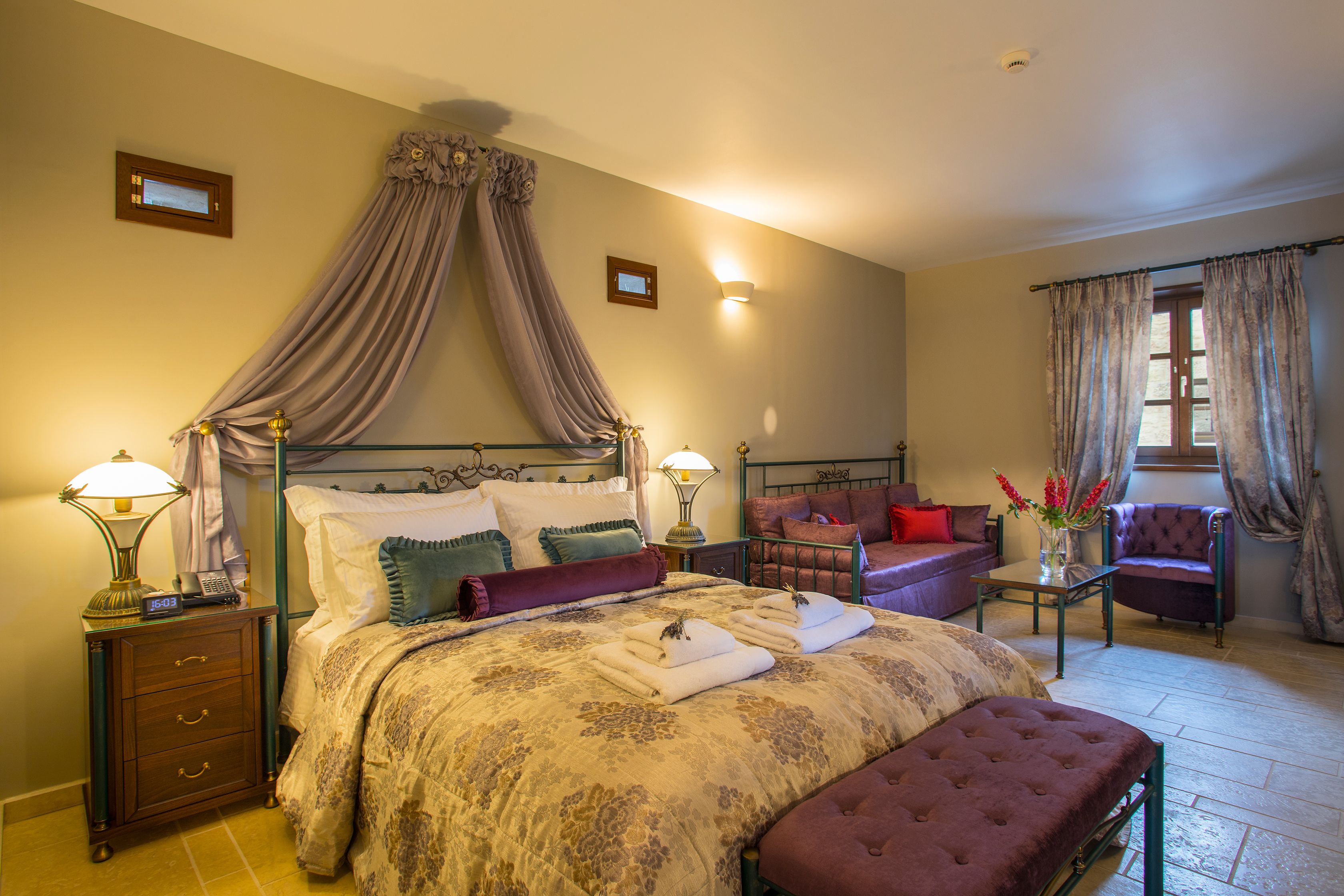 Un dormitorio bien iluminado con una cama con dosel, una mesita de noche con una lámpara, una mesilla con un jarrón de flores y una zona de estar con una silla y un sofá.