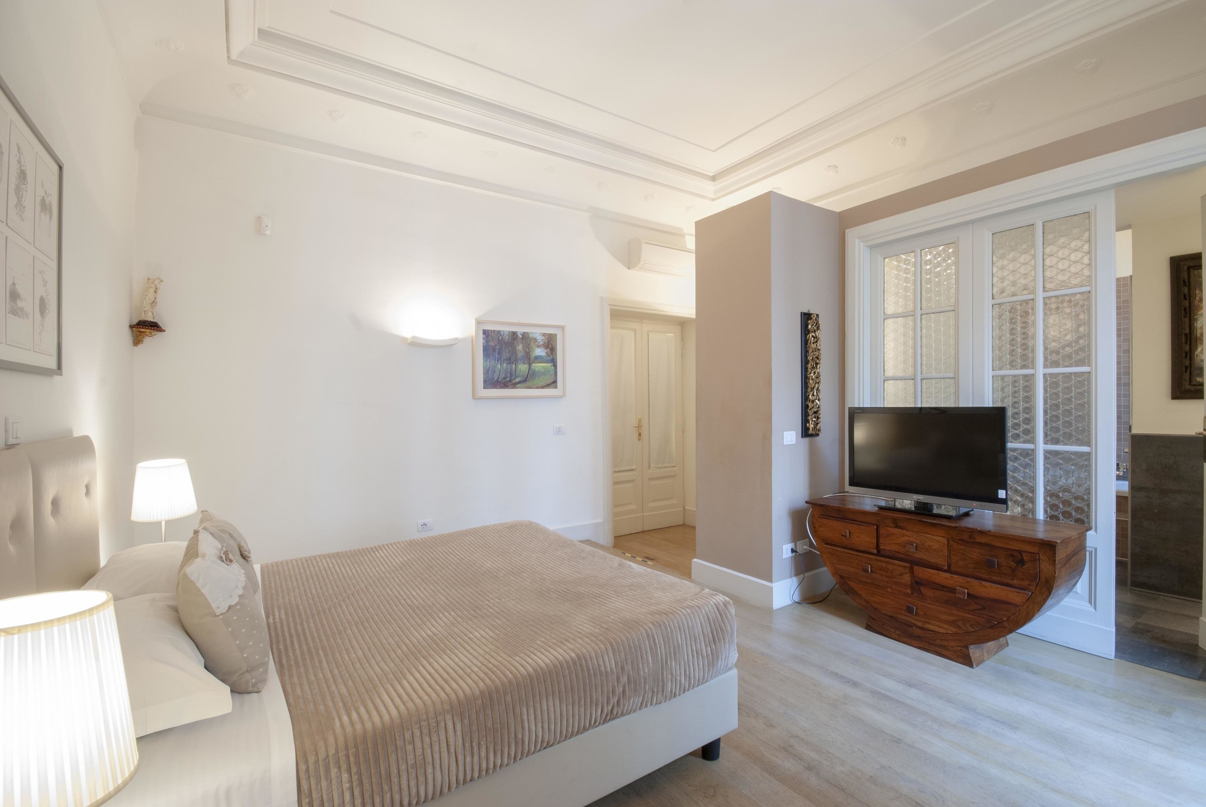A bedroom with a bed, a TV on a wooden dresser, and a painting on the wall.