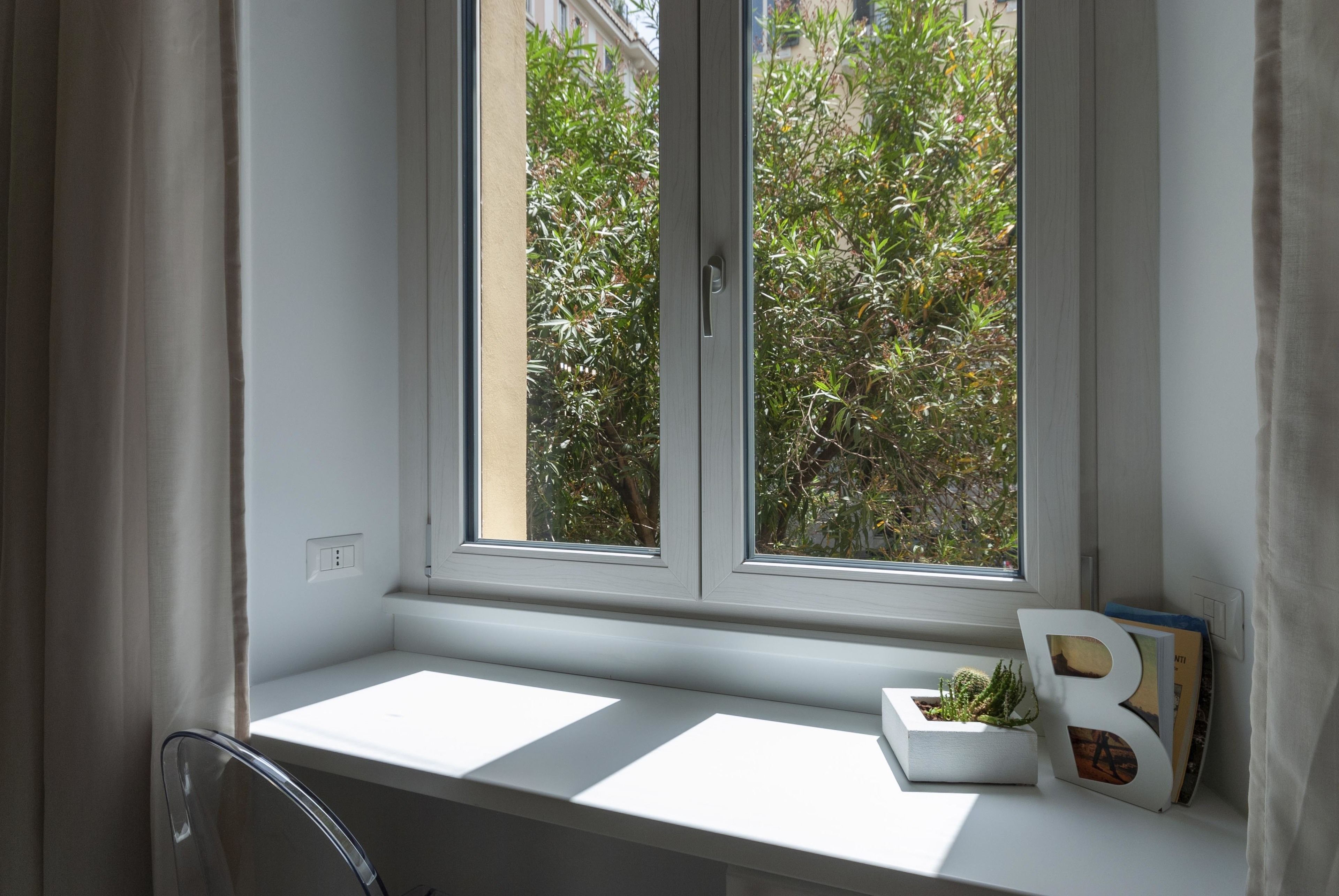 A window with a view of greenery outside, a white shelf with a plant and a letter 'B' decoration, and a chair to the left.