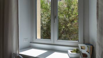 A window with a view of greenery outside, a white shelf with a plant and a letter 'B' decoration, and a chair to the left.