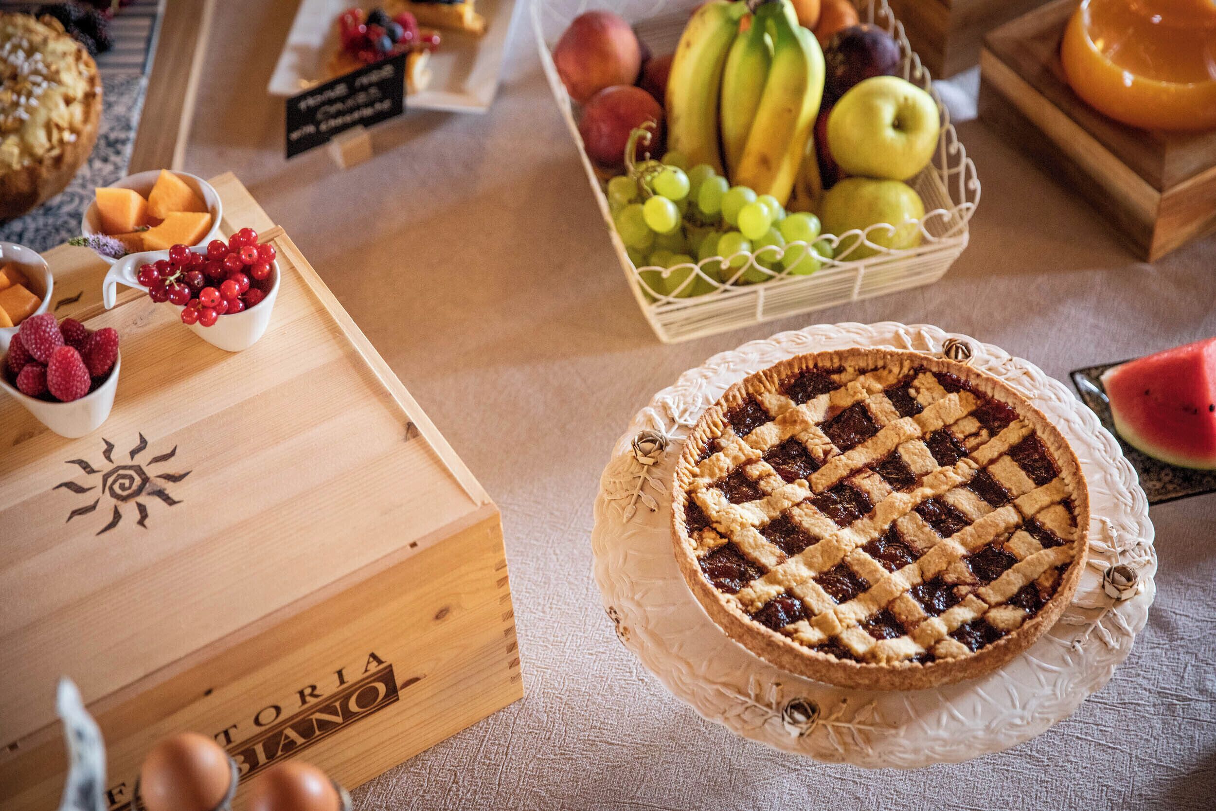 A wooden crate labeled "FONTAINA BIANO" with a sun design, a pie with a lattice crust, and a basket of fruits including grapes, apples, and bananas.