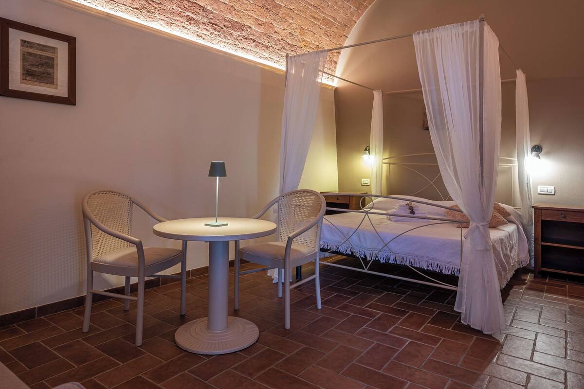 A bedroom with a bed, a canopy, two chairs, a small table, and a lamp.