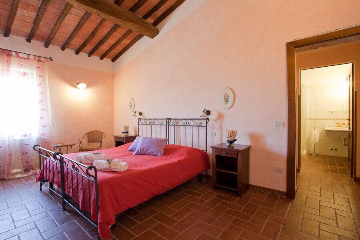 A bedroom with a red bed, a wooden ceiling, a chair, a nightstand, and a bathroom visible through an open door.
