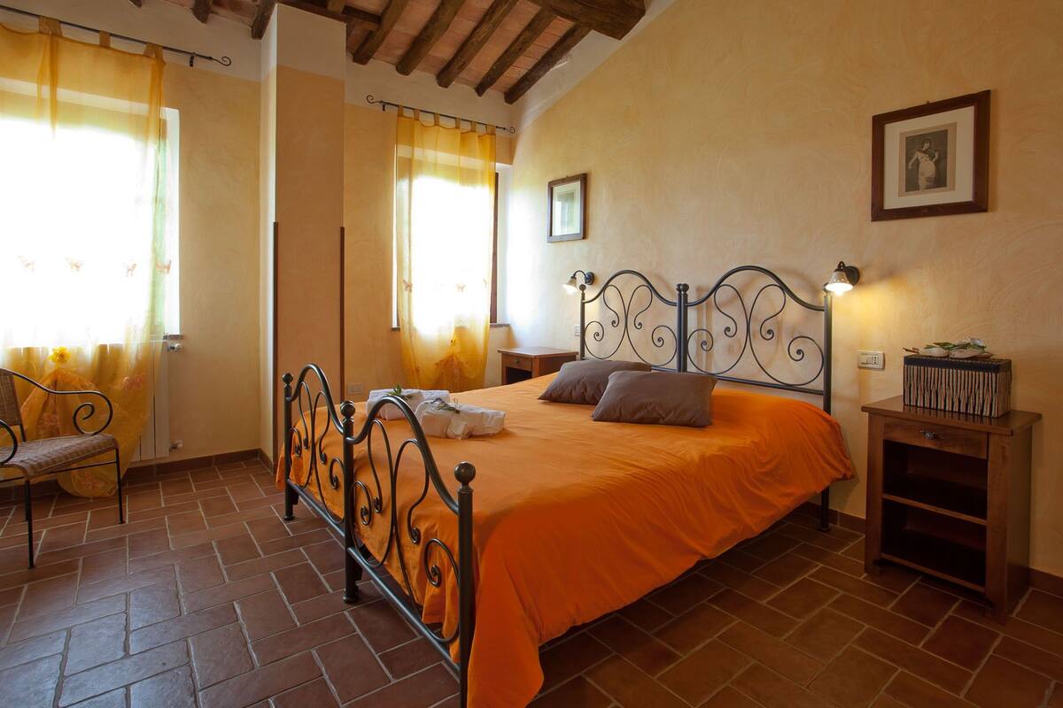 A bedroom with a metal bed frame, an orange bedspread, a chair, a nightstand, and a lamp.
