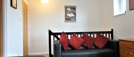 A room with a black leather sofa with red cushions, a wooden dresser, a framed picture on the wall, and a window with a view.