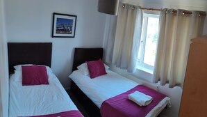 Une chambre d’hôtel avec deux lits simples, chacun recouvert d’une courtepointe blanche et rose, doté d’une tête de lit marron et d’un oreiller blanc. Un tableau encadré est accroché au mur, il y a une fenêtre avec des rideaux et un radiateur sous la fenêtre.