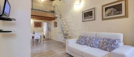 A spacious living area with a white sofa, a dining table with chairs, a staircase, and a ceiling with exposed beams.
