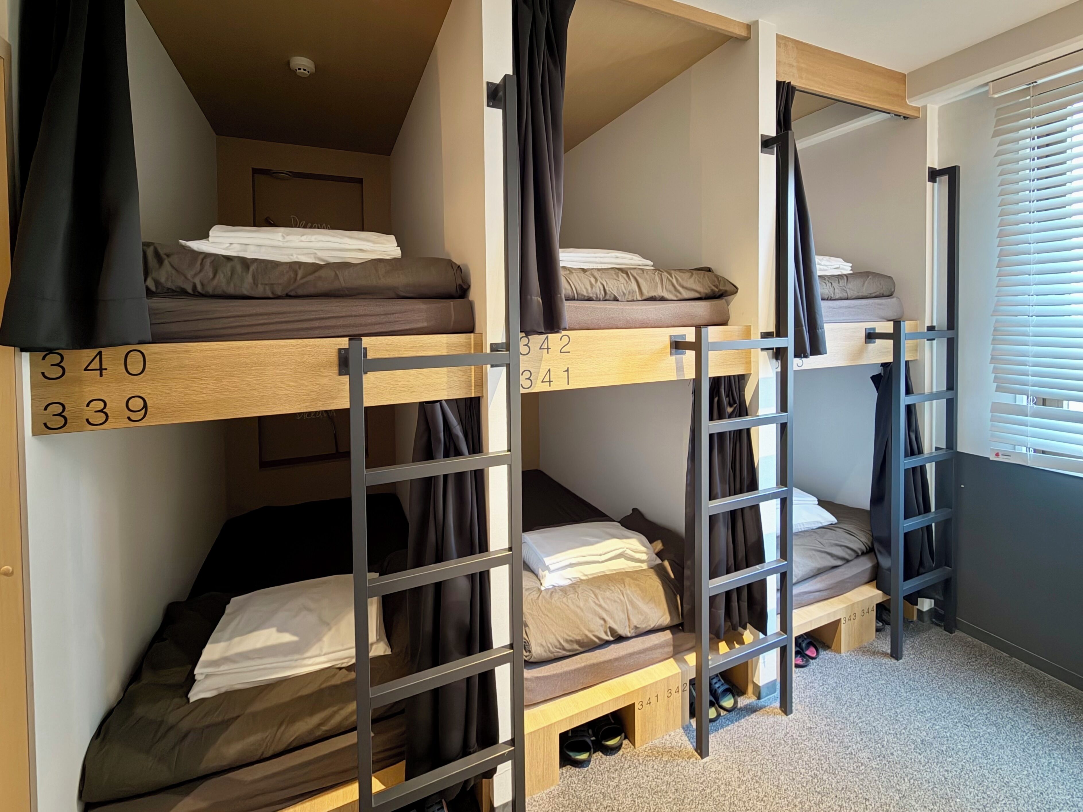 A dormitory room with bunk beds, numbered beds, and a window with blinds.