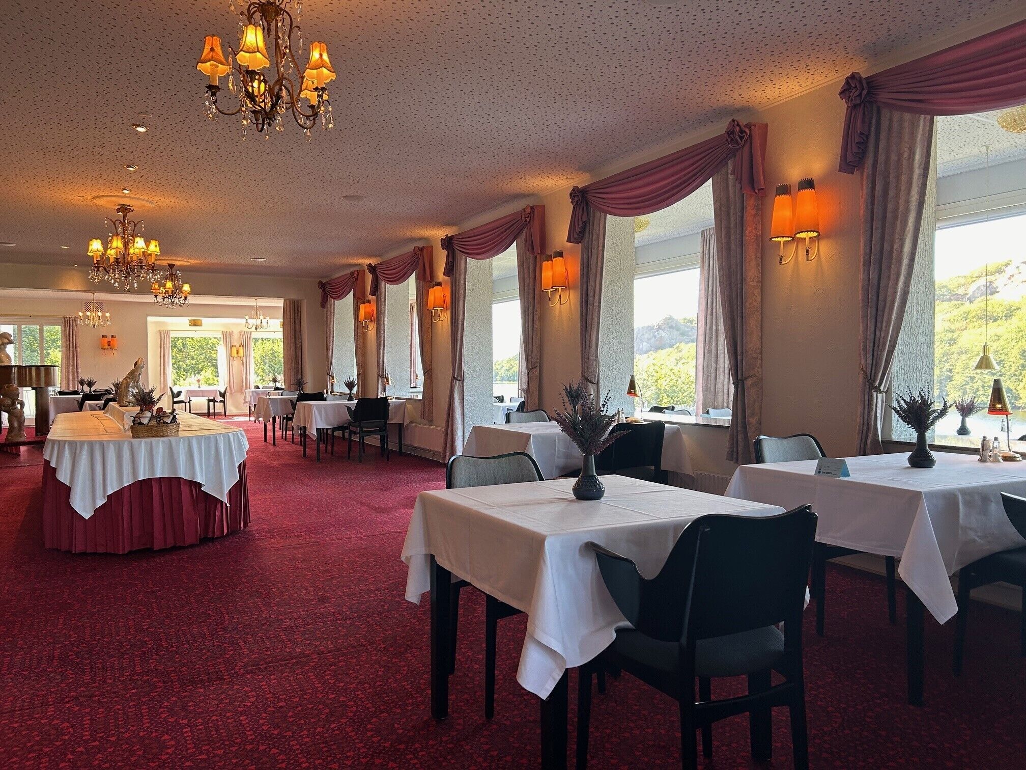 Un restaurante con varias mesas, candelabros y amplias ventanas con cortinas.
