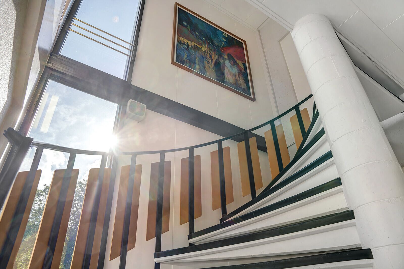 A staircase with a glass railing, a painting on the wall, and a view of the sky through the windows.