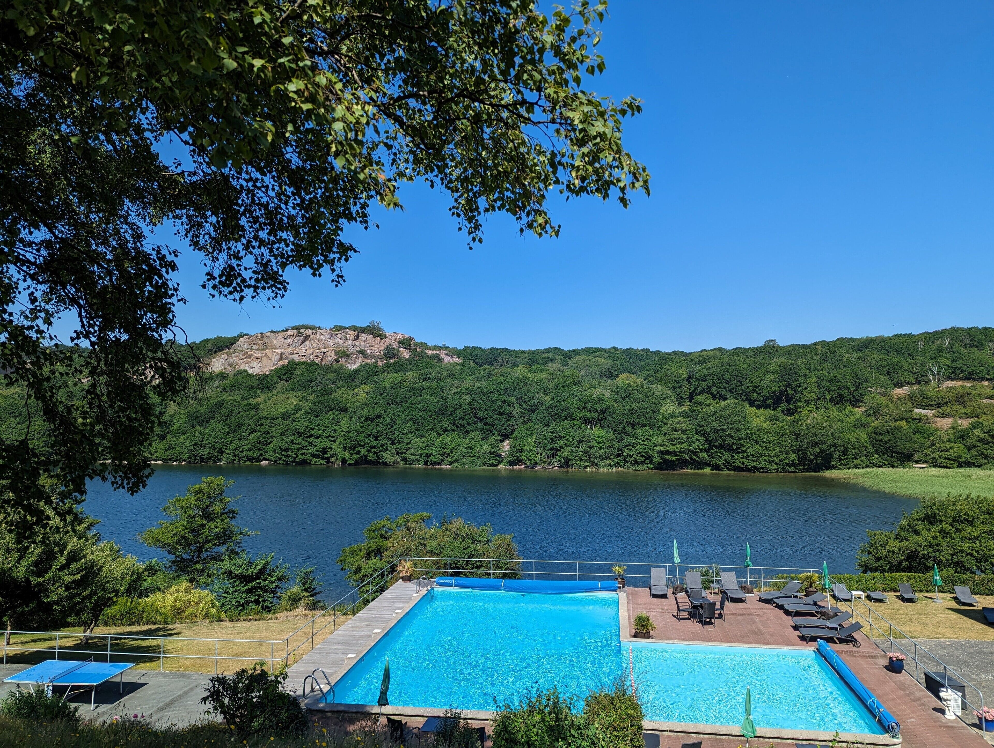 En swimmingpool med liggestole og parasoller, omgivet af frodig vegetation og en sø med en klippe bakke i baggrunden.