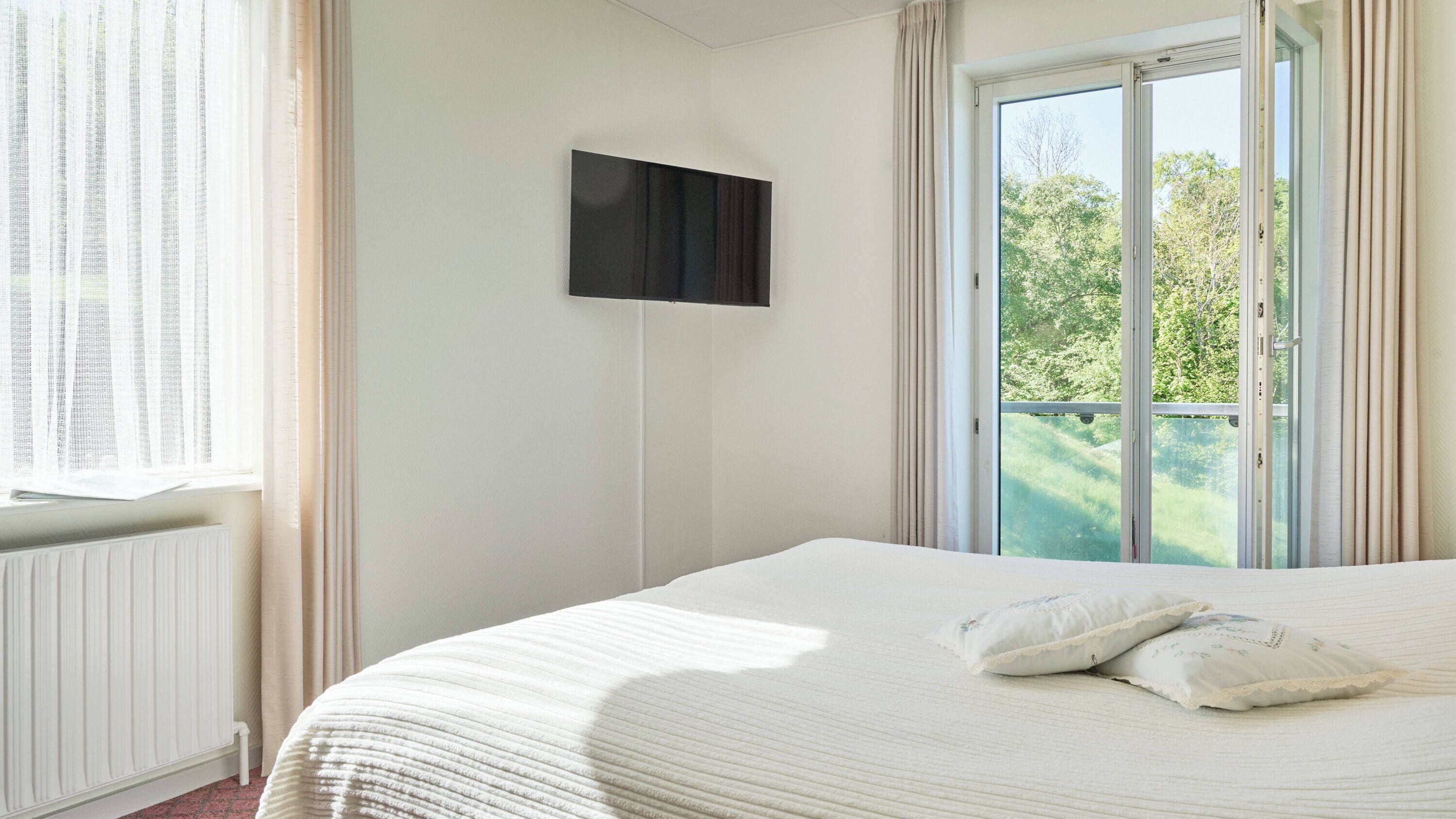 A bedroom with a large bed, a flat-screen TV mounted on the wall, and a sliding glass door leading to a balcony.
