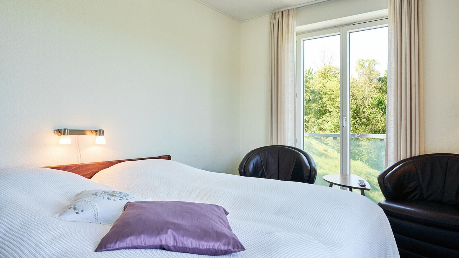 Hotellihuone, jossa on sänky, tuoli, pieni pöytä ja ikkuna, josta avautuu näkymä vehreään maisemaan.
