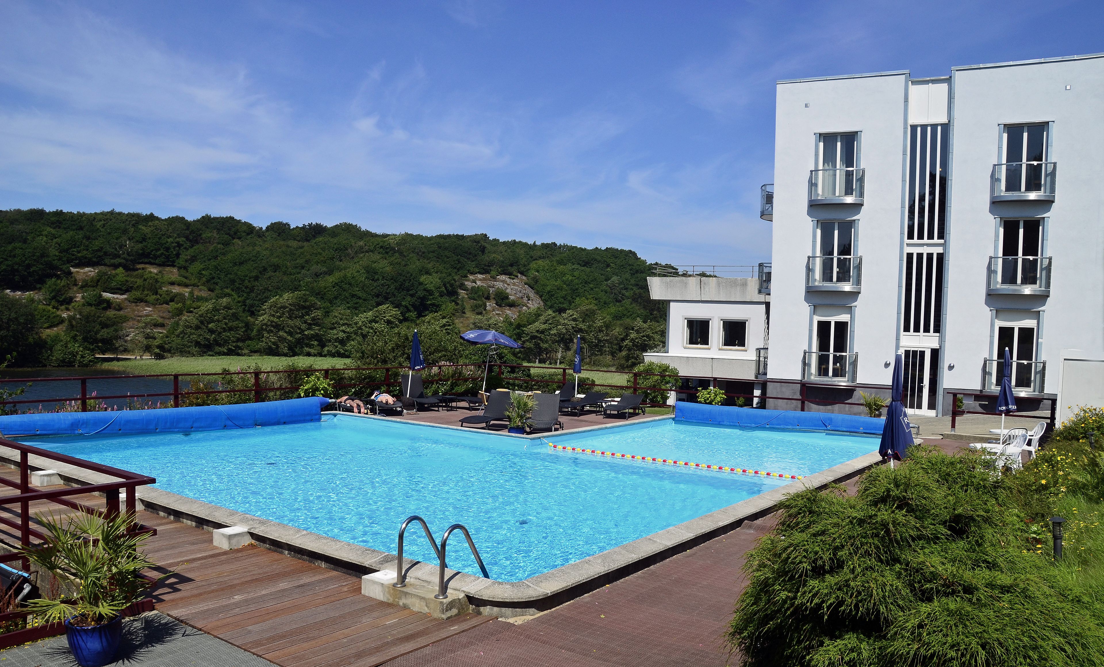 Una piscina con sillas de descanso y sombrillas, rodeada por una terraza de madera y un edificio con balcones.