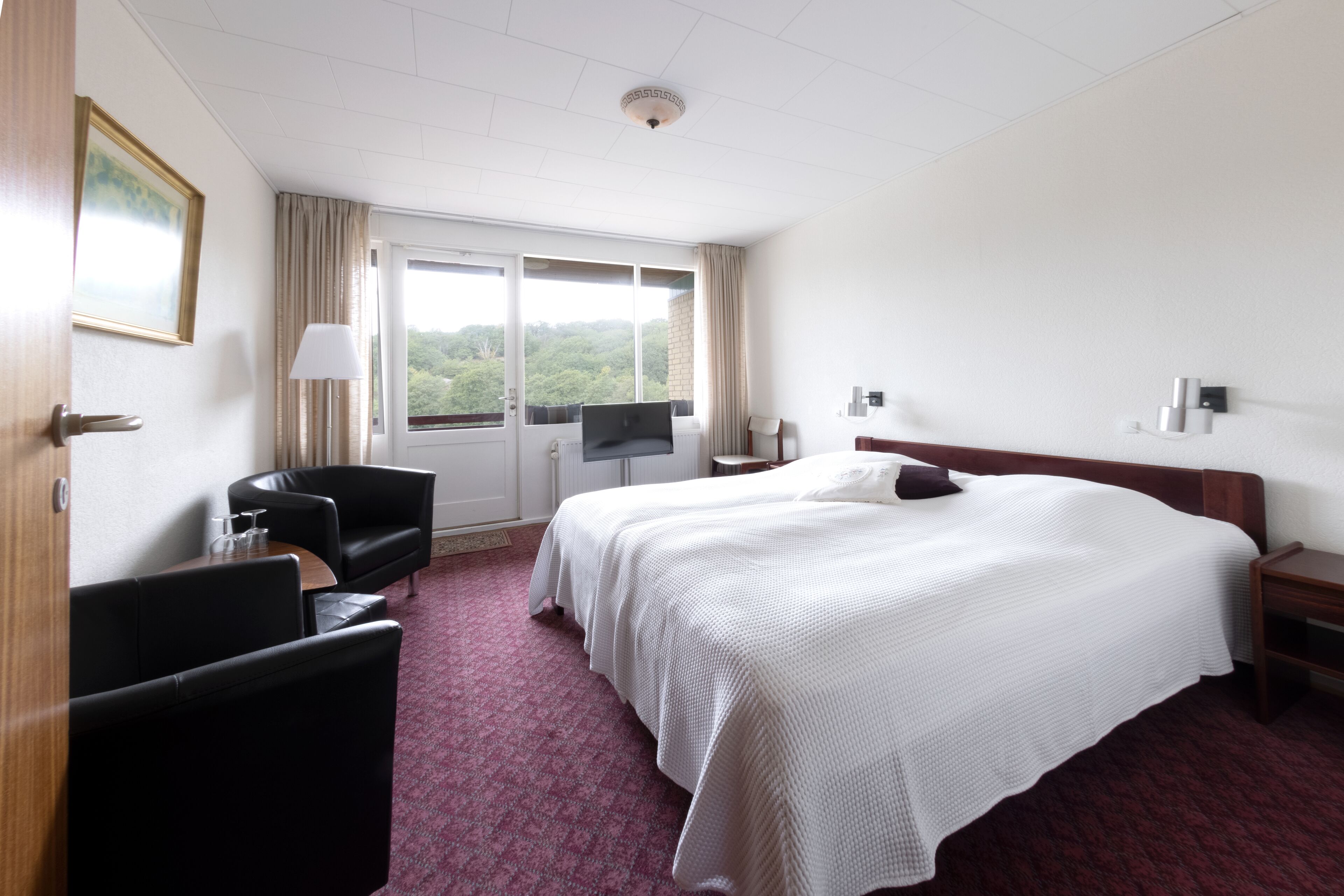 A hotel room with a large bed, a desk, a chair, and a window with a view of greenery.