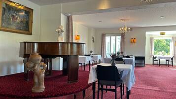 A dining area with a red carpet, tables, chairs, a grand piano, and a chandelier.
