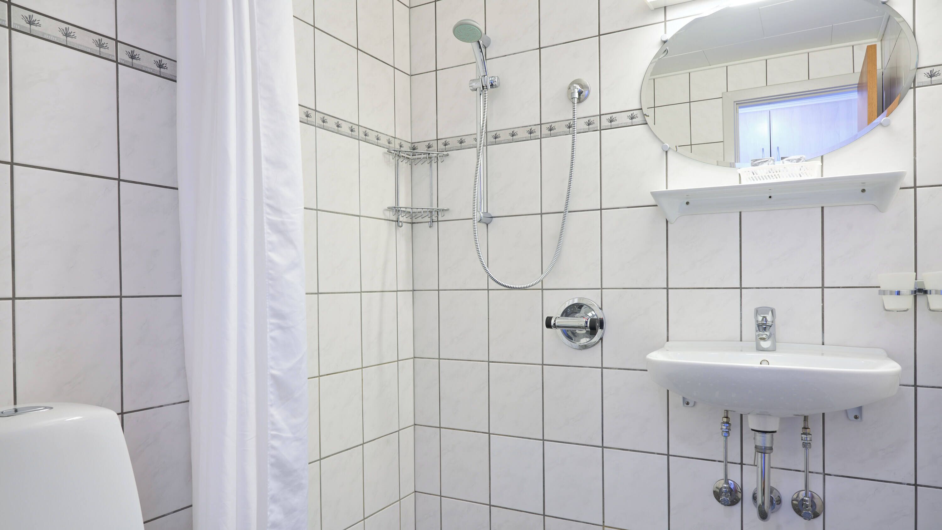 Une salle de bain avec des murs carrelés de blanc, une douche avec un pommeau de douche amovible, un lavabo avec un miroir au-dessus et des toilettes.