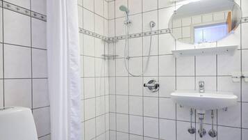 Une salle de bain avec des murs carrelés de blanc, une douche avec un pommeau de douche amovible, un lavabo avec un miroir au-dessus et des toilettes.
