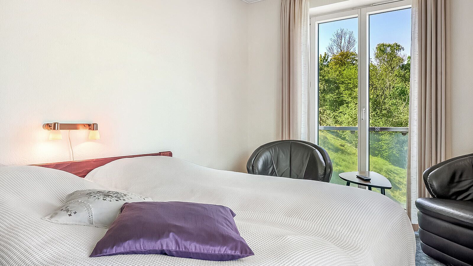 A hotel room with a bed, a chair, a small table, and a balcony with a view of greenery.