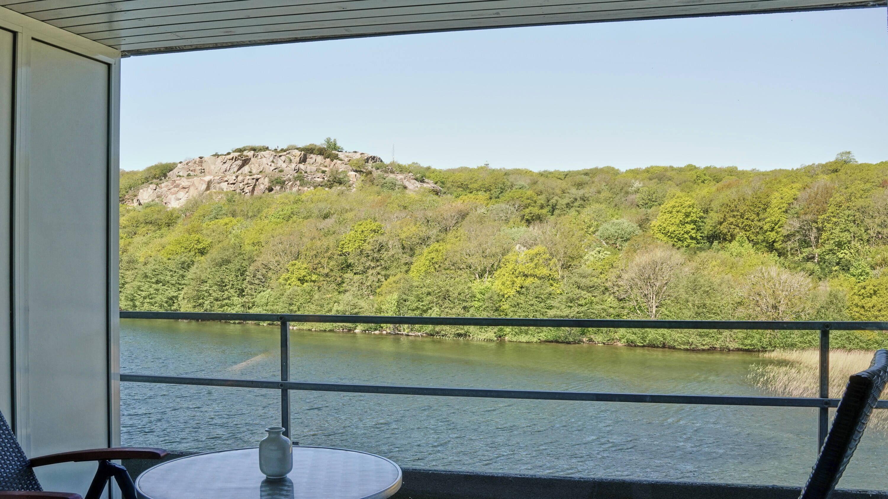 Balcón con una mesa y un jarrón, con vistas a un río, una colina rocosa y abundante vegetación al fondo.