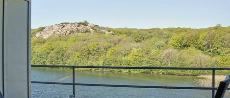 Balcón con una mesa y un jarrón, con vistas a un río, una colina rocosa y abundante vegetación al fondo.