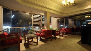 Un vestíbulo de hotel con techo alto, ventanas amplias y un área de descanso con sillas rojas y una mesa de madera.