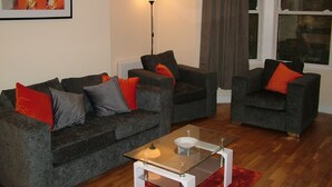 Ein Wohnzimmer mit zwei grauen Sofas, roten und grauen Kissen, einem Glastisch, einer Stehlampe und einem Fenster mit Vorhängen.