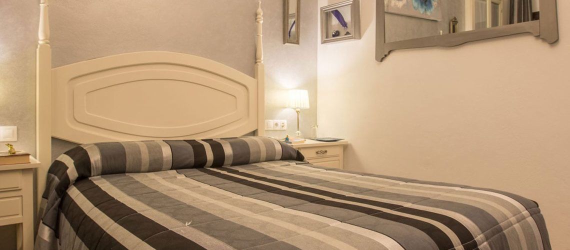 A neatly made bed with a striped bedspread and a matching pillow, a bedside table with a lamp, and a wall-mounted mirror.